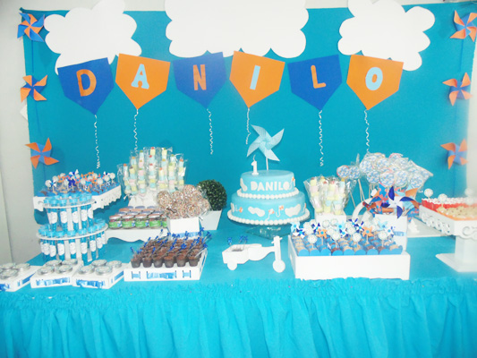 Imagem mostra uma festa de aniversário brasileira com uma mesa decorada com panos azul, bolo e docinhos com o tema de catavento