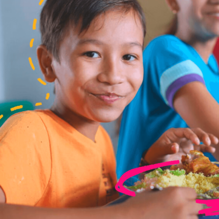 Imagem de capa para matéria sobre a política de merenda escolar mostra um menino pardo de camiseta laranja sentado em uma mesa e comendo um prato de comida