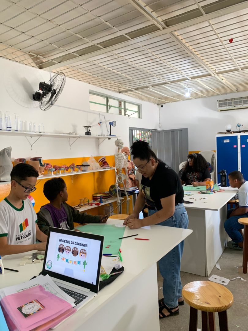 Imagem mostra um laboratório de escola, um computador em uma mesa, alunos sentados ao redor e a professora orientando.