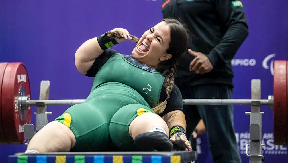 Imagem mostra a atleta da paralimpíadas, Mariana Dandrea, uma mulher branca, com naninsmo, vestindo uniforme da seleção brasileira e comemorando após a prova de halterofilismo