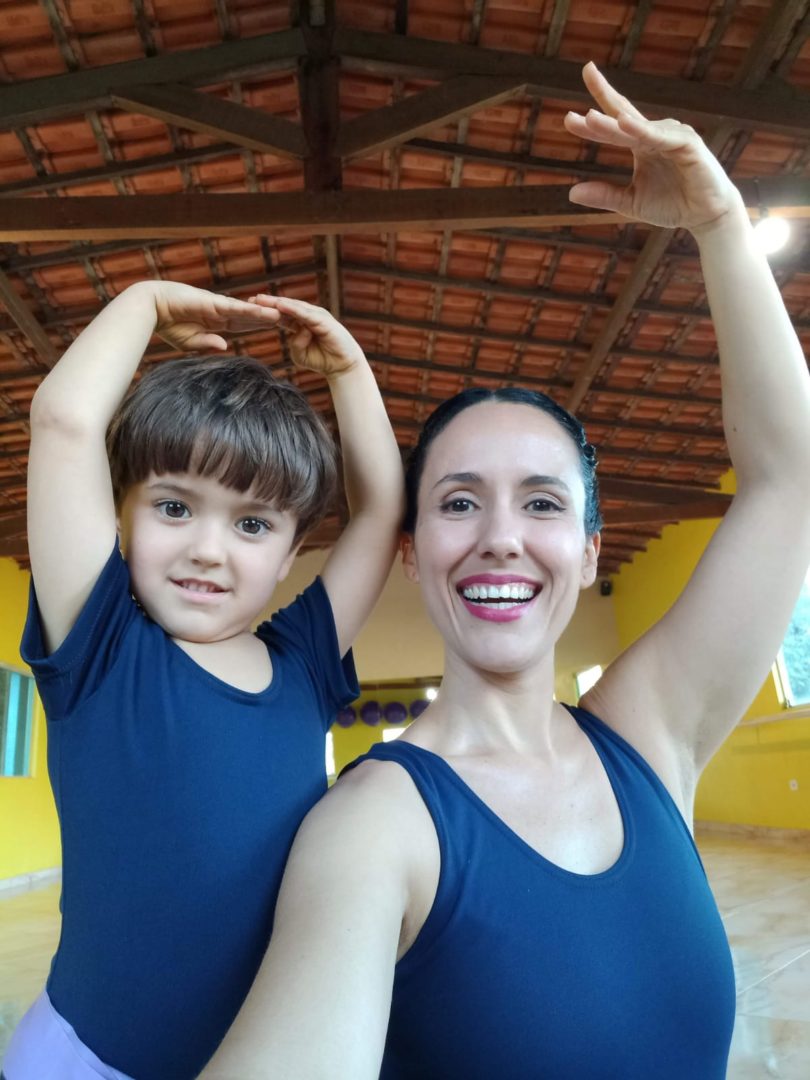 imagem mostra uma mulher branca com roupa de balé com os braços levantados ao lado de um menino branco, de cabelos curtos fazendo a mesma pose