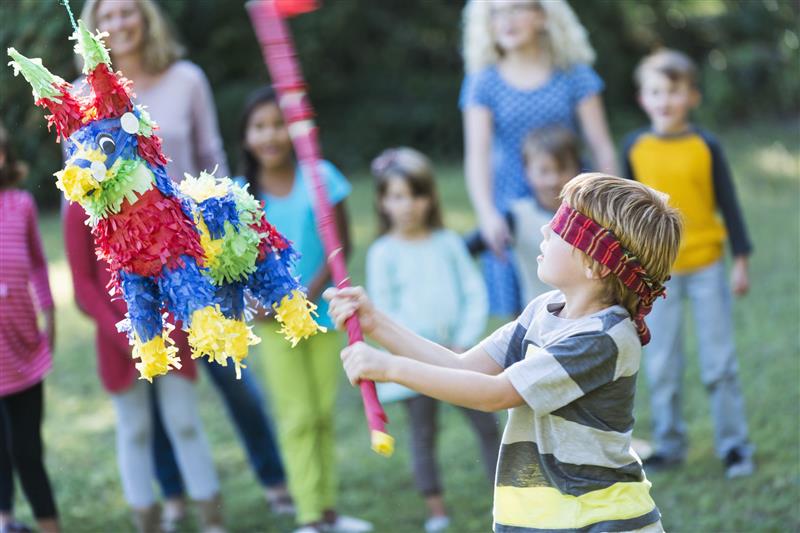 Imagem mostra uma das festas de aniversário pelo mundo. Um menino branco, de olhos vendados, tentando quebrar uma piñata colorida com um bastão de madeira.