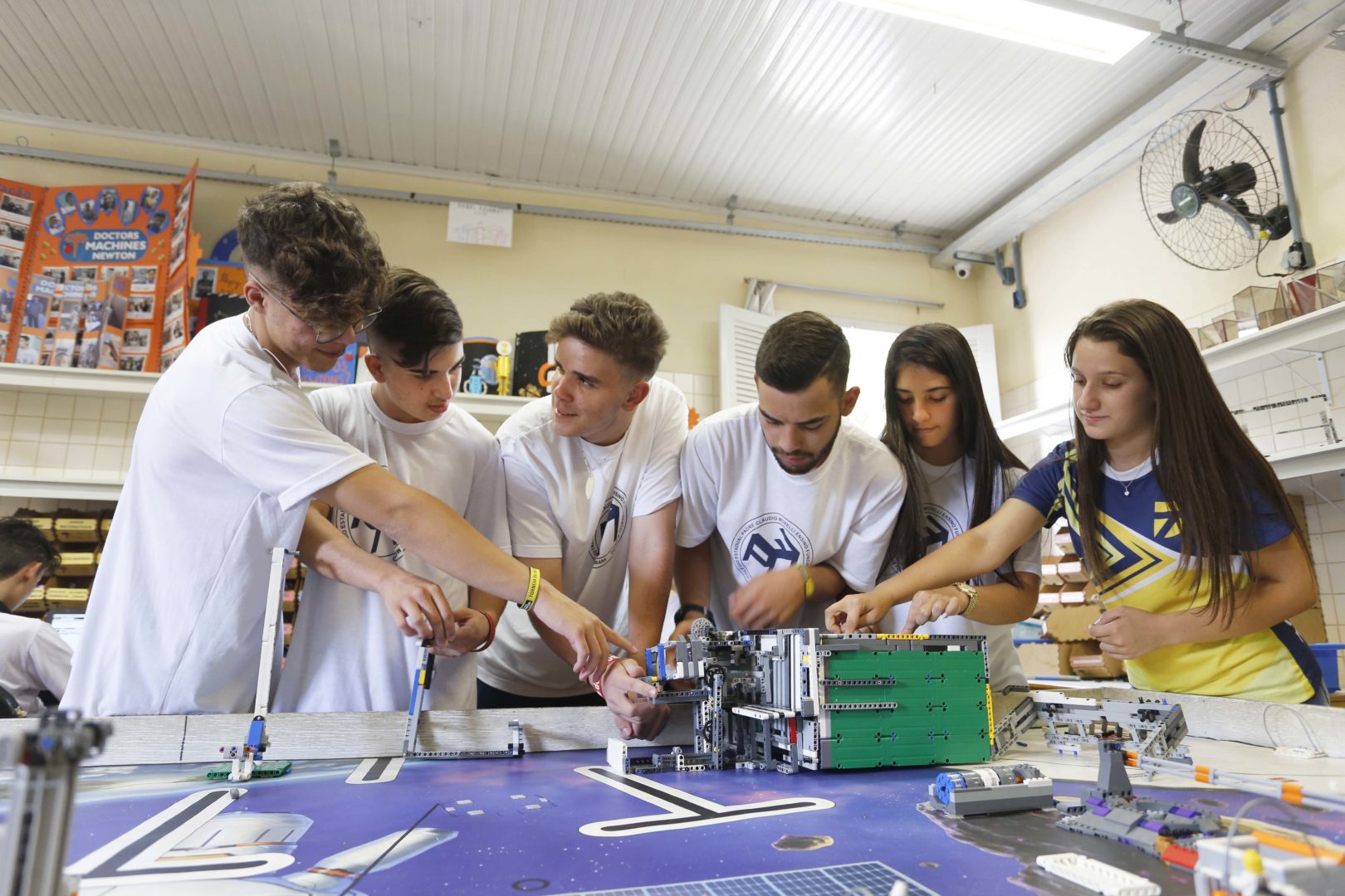 Imagem mostra cinco adolescentes em uma aula de robótica educacional no Paraná. Todos estão em pé ao redor de uma mesa onde há um protótipo de robô.