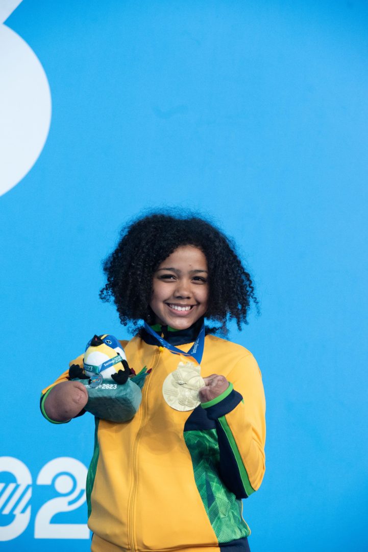 Imagem mostra atleta da paralimpíadas, Alessandra, uma menina negra, de cabelos cacheados, usando um casaco com cores da bandeira do Brasil e uma medalha de ouro. 