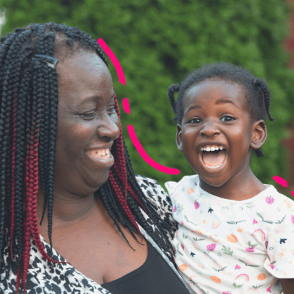 Uma mulher negra de tranças segura no colo uma menina também negra que usa uma camiseta branca com estampa colorida.