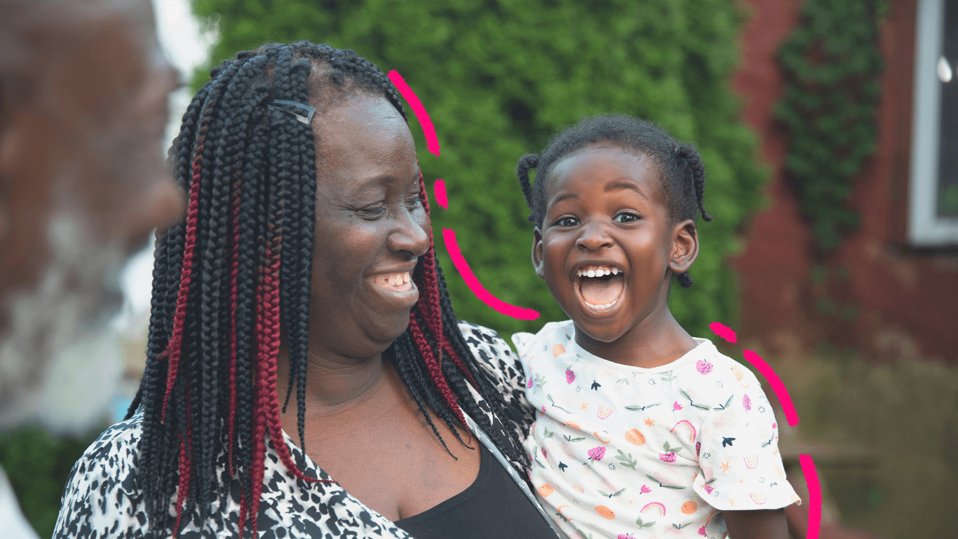 Uma mulher negra de tranças segura no colo uma menina também negra que usa uma camiseta branca com estampa colorida.