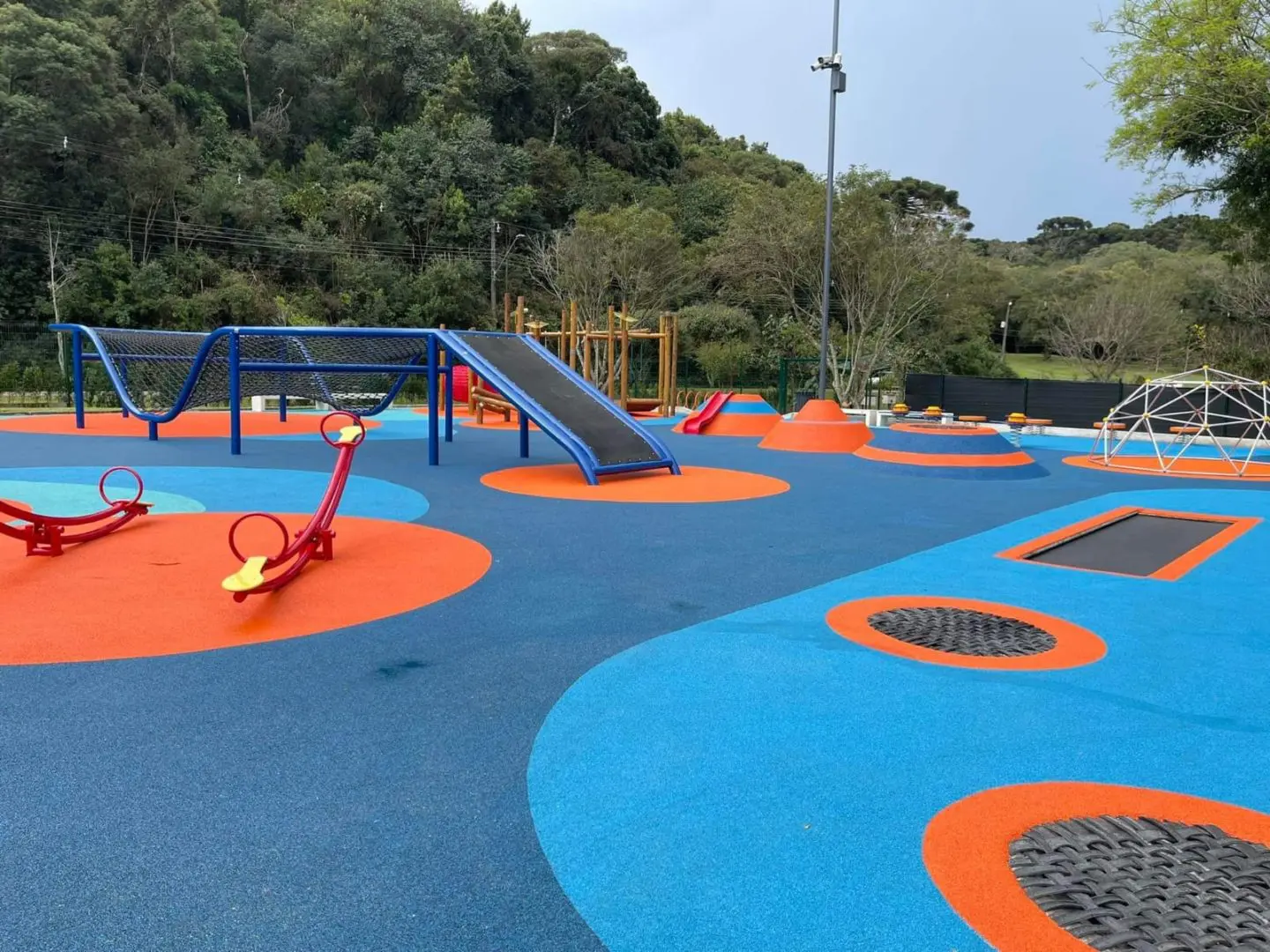 Imagem de parques inclusivos mostra o parquinho do Birigui com o chão em azul e laranja, brinquedos de ferro coloridos e crianças brincando no espaço.