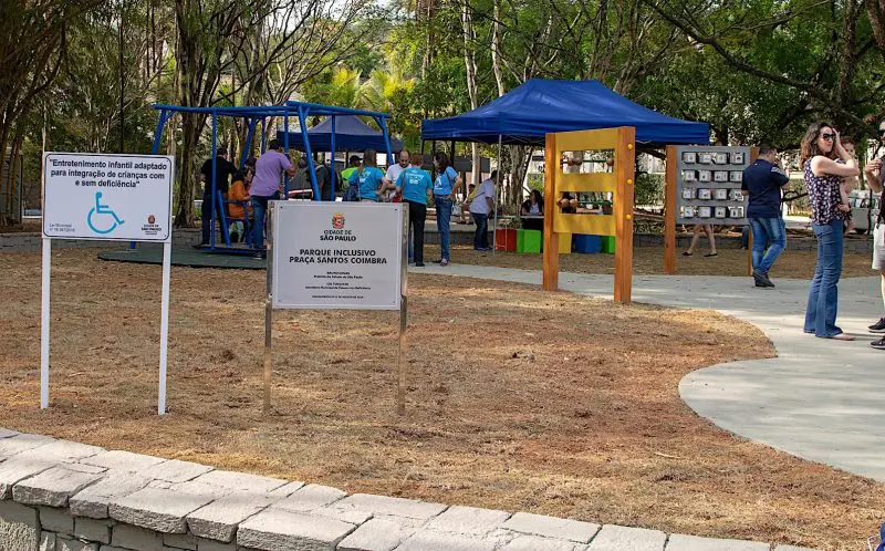 Imagem de parquinhos inclusivos mostra o espaço da praça Santos Coimbra, com brinquedos coloridos e pessoas no local. 