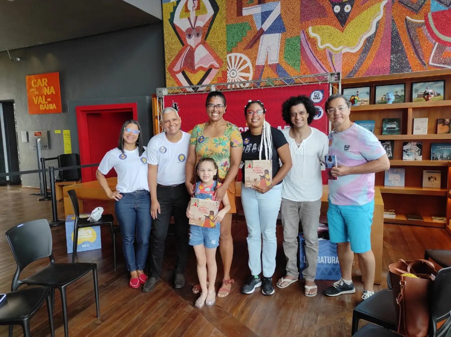 Imagem mostra o clube de leitura Lendo a Bahia com pessoas segurando livros nas mães e um acriança, menina branca de cabelos cacheados, no centro do grupo.