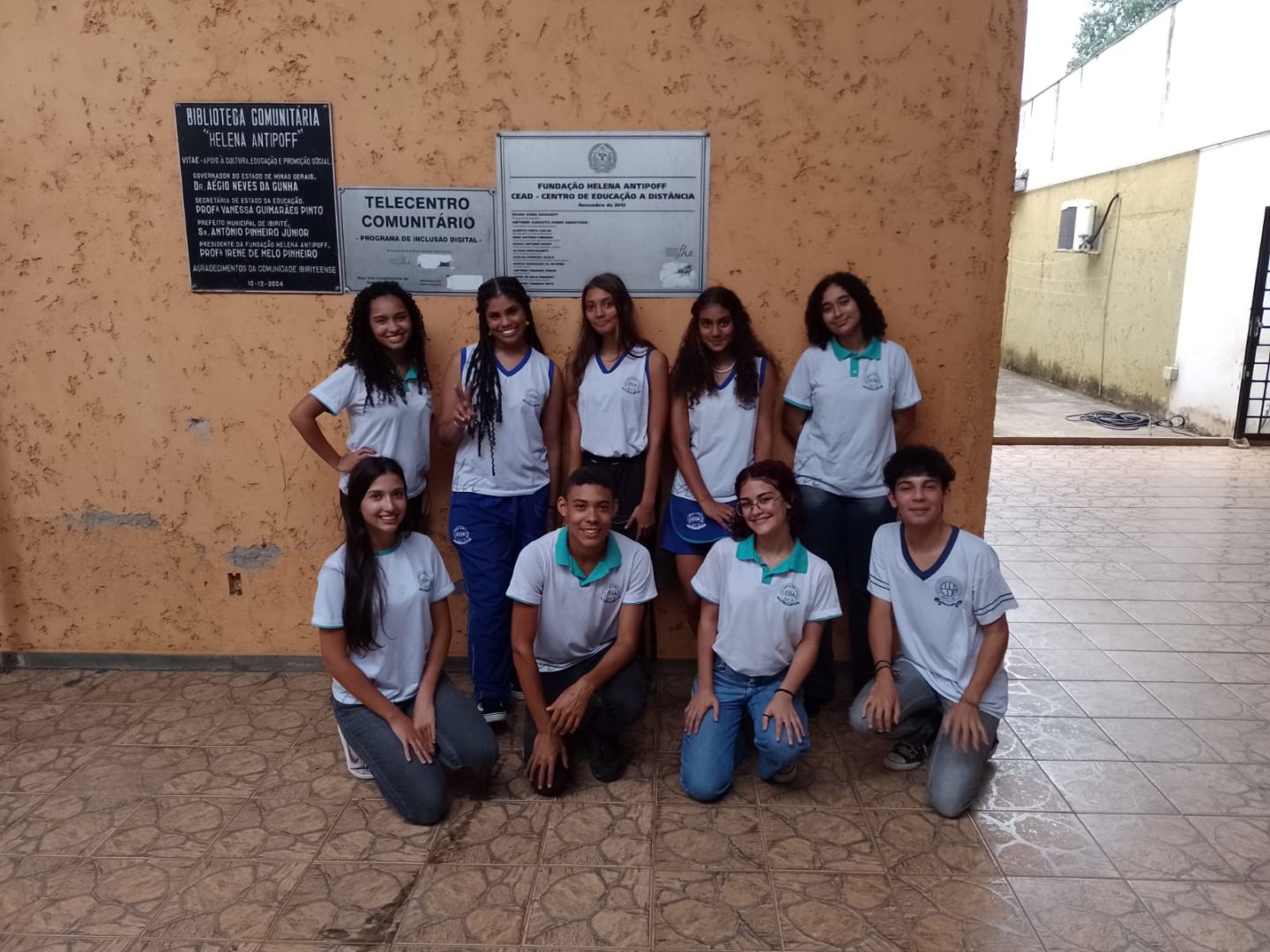 Imagem de estudantes do clube de leitura mostra nove adolescentes vestindo uniforme da escola e posando para a foto em frente a uma biblioteca 