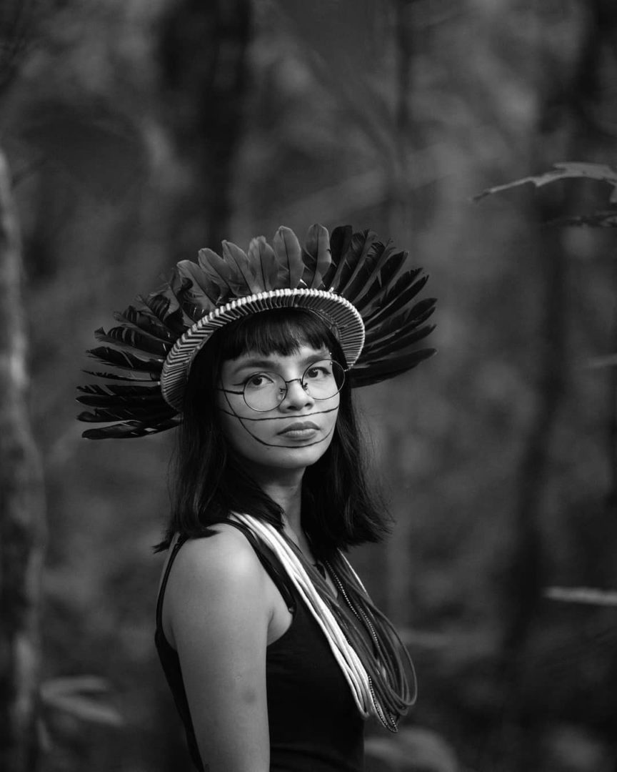 Imagem mostra uma fotografia de Txai Suruí, uma mulher indígena, de cabelos lisos e negros, rosto com pinturas e cocar de penas.