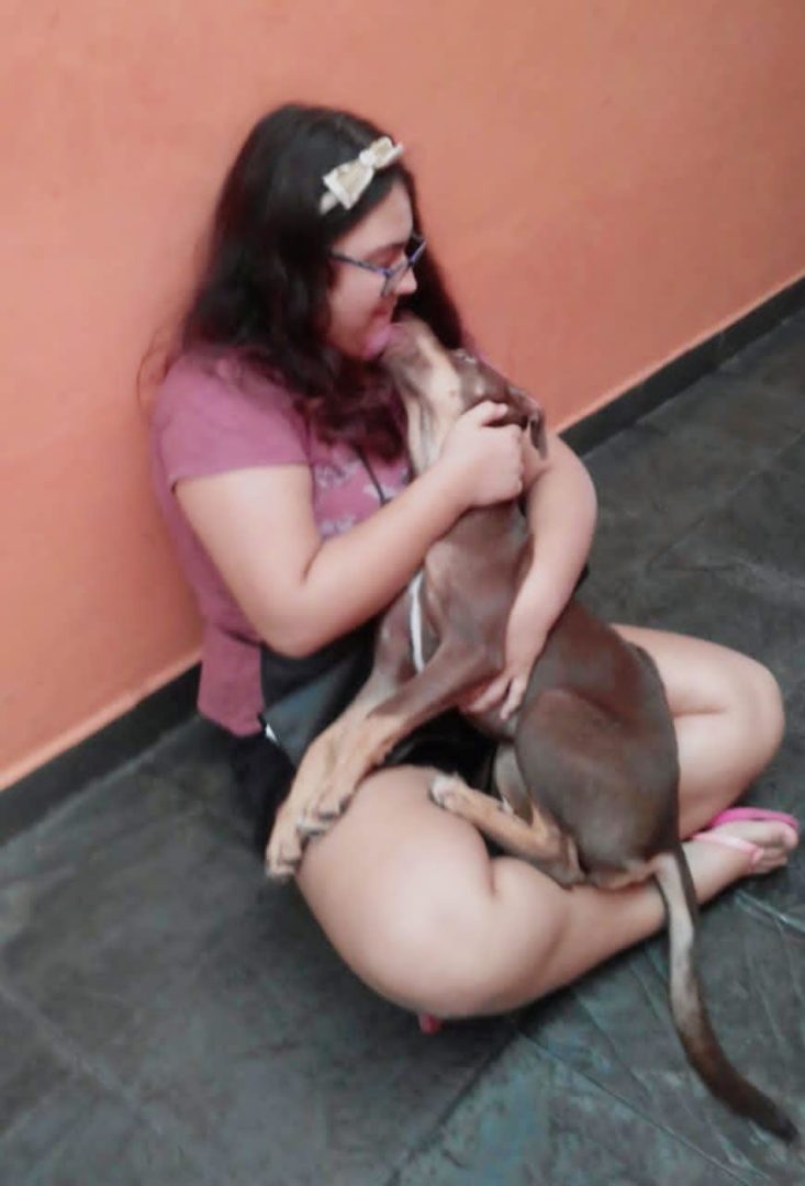 Imagemmostra Giovana, uma adoelscente branca, de cxabelos ondulados castanhos. Ela está sentada no chão de uma casa e brincando com sua cachorrinha. 