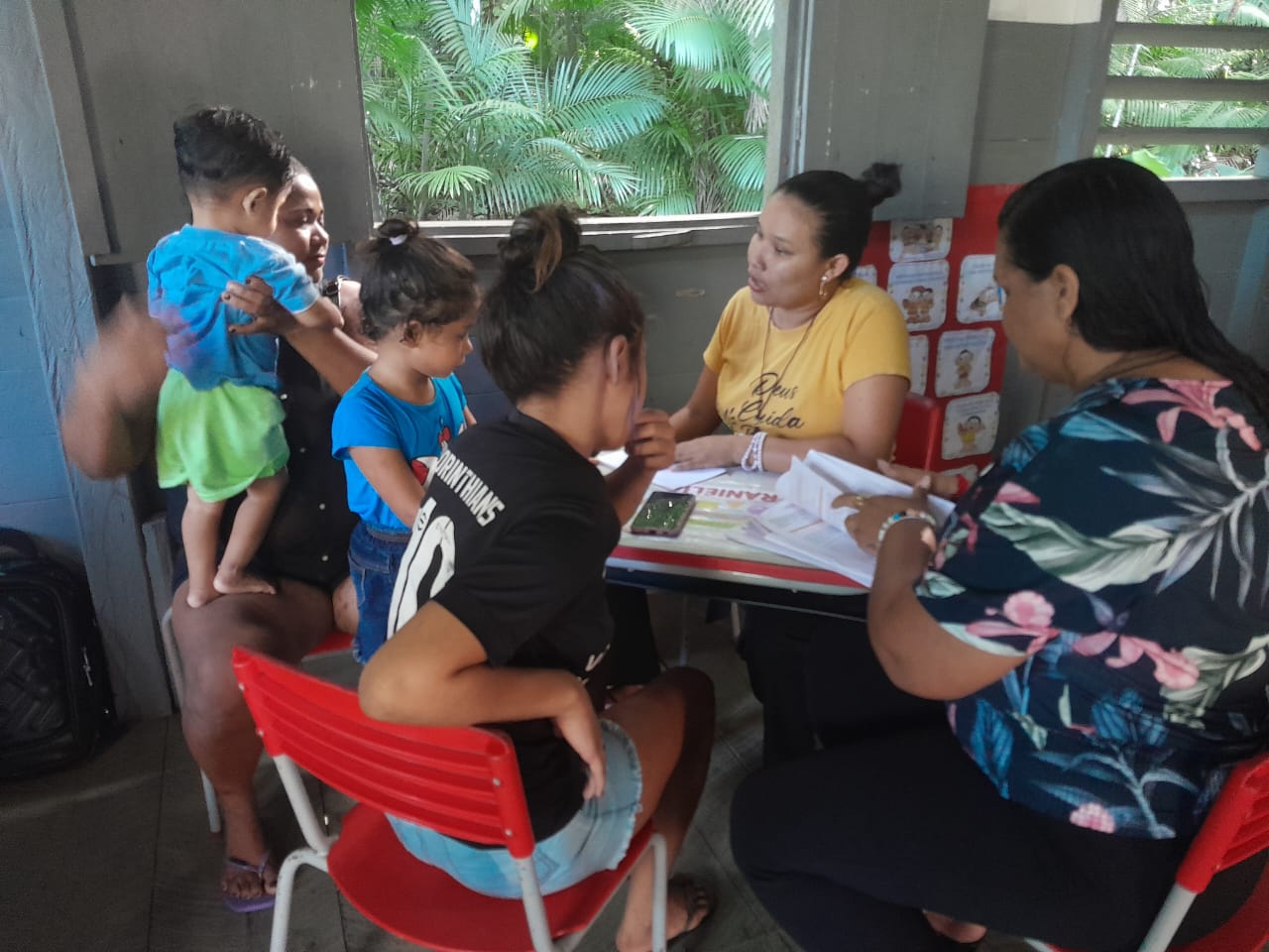 Imagem mostra uma reunião entre escolas e famílias em uma sala com carteira svermelhas, adultos e cianás sentados e a paisagem da floresta pela janela.