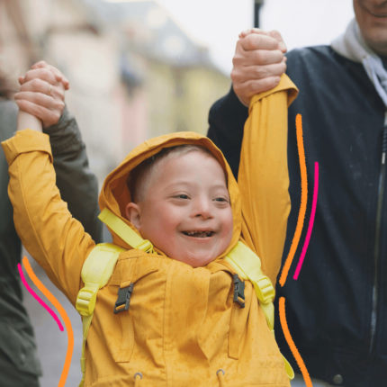 Imagem de capa para matéria sobre como as infâncias transformam os adultos mostra um menino branco, usando casaco amarelo, sorrindo e sendo carregado pelos braços por dois adultos.