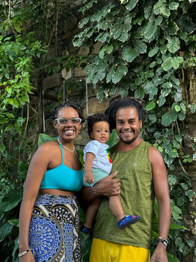 Imagem de personagem sobre nome de bebê mostra a família de Dendê. À esquerda, há uma mulher negra com tranças no cabelo. Ela usa óculos e veste um top azul, com uma saia azul de estampas africanas. À direita, ao seu lado, está um homem negro com dreads no cabelo. Ele veste uma camiseta regata de cor verde musgo e uma bermuda amarela. Ele está segurando nos braços uma criança negra. 