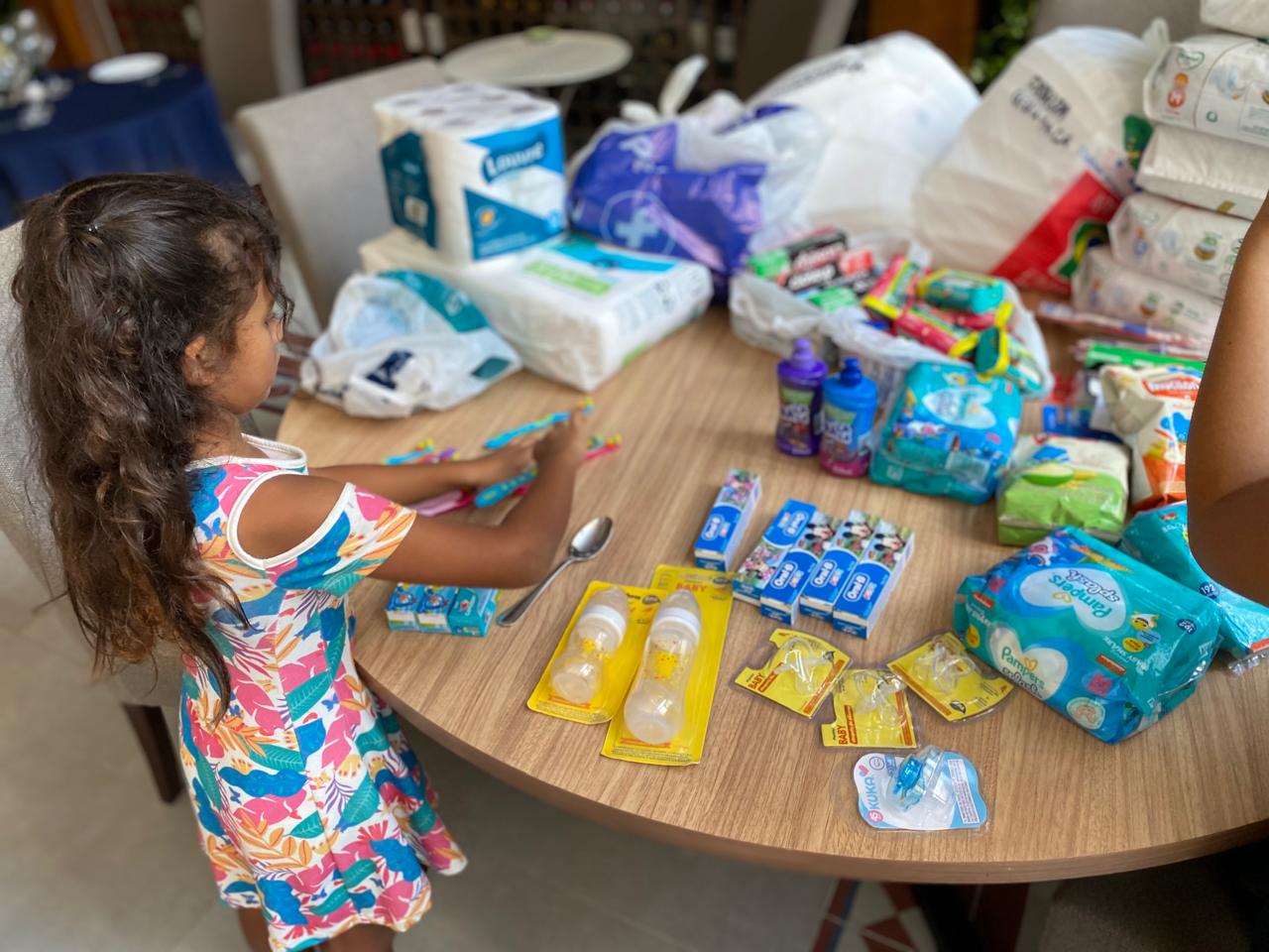 Imagem mostra Joana, uma menina negra, de cabelos longos ondulados e vestido florido. Ela está de frente para uma mesa organizando produtos de higiene para doação.