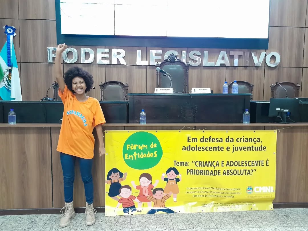 Imagem mostra Pérola, uma adolescente negra, de cabelos médios cacheados, vestindo calça jeans e camiseta laranja. Ela levanta o braço direito e posa para a foto ao lado de uma faixa sobre os direitos das crianças dentro de um prédio público.