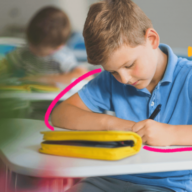 Imagem de capa para matéria sobre pressão por boas notas na escola mostra um menino branco, de cabelo scurtos loiro, vestindo camisa azul. Ele está sentado em uma carteira, em sala de aula e escrevendo em um caderno.