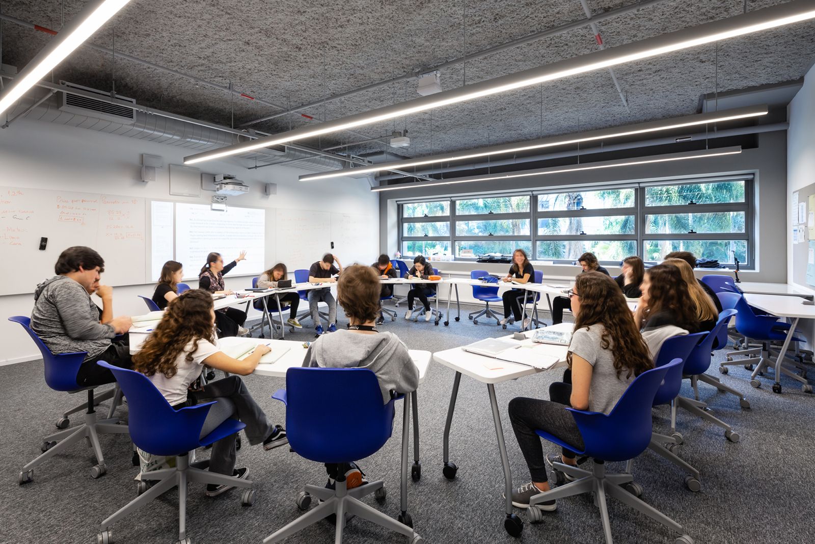 Imagem mostra uma sala de aula com estudantes sentados em carteiras dispostas em círculo. 