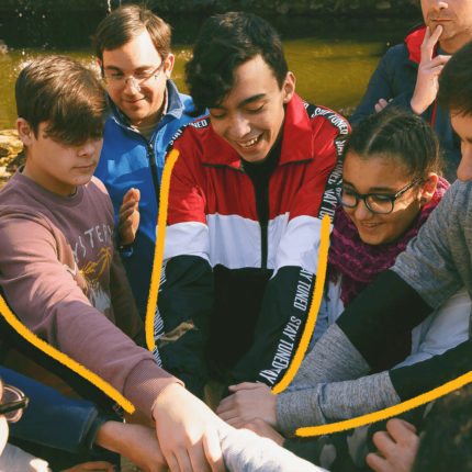 Imagem mostra um grupo de adolescentes vestindo roupas coloridas e unindo as mãos em uma roda.