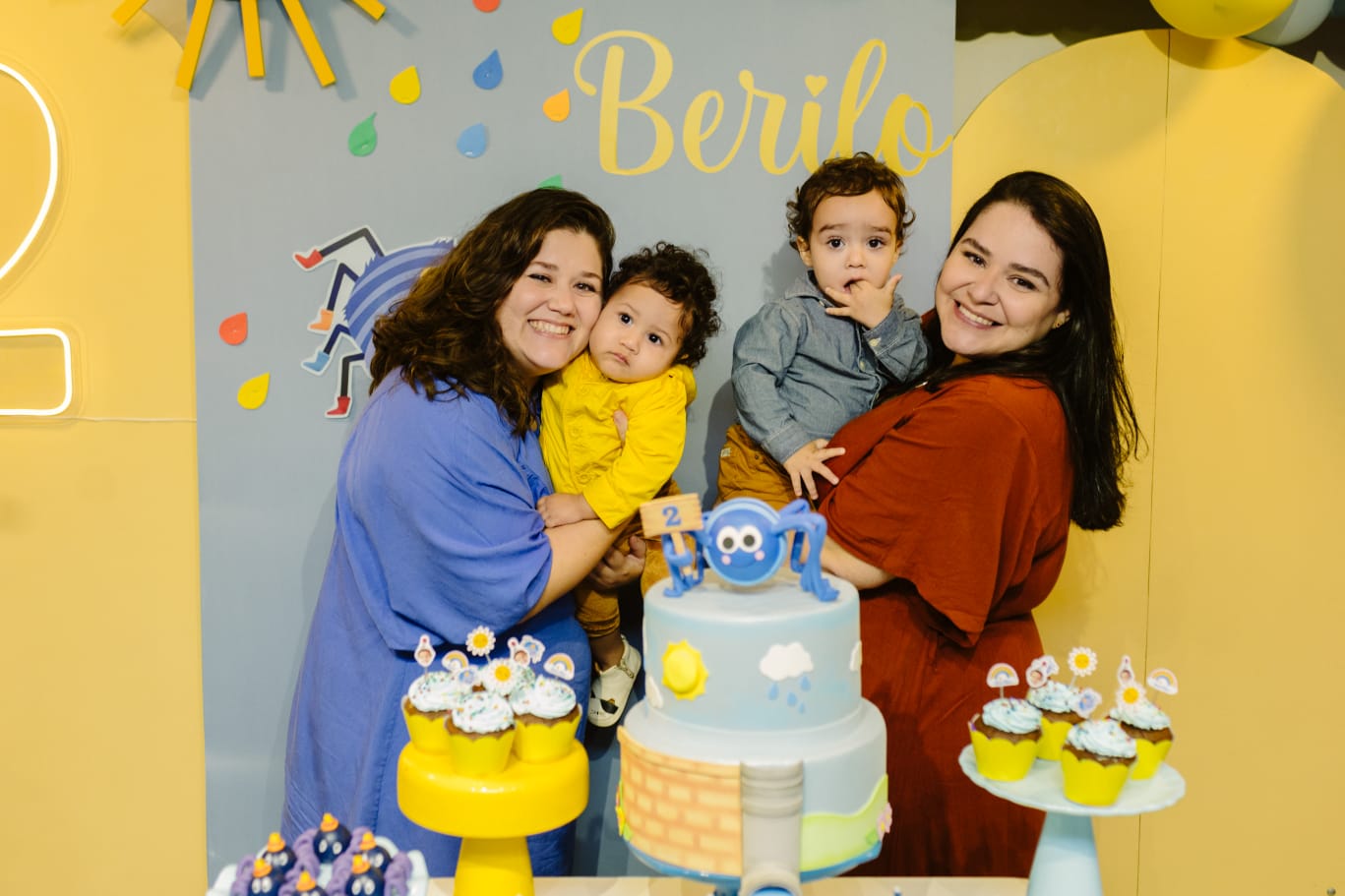 Imagem mostra uma festa de aniversário, onde uma mulher branca, de cabelos médios castanhos, está de vestido azul segurando um bebê no colo. Ao lado, uma outra mulher branca, de cabelos longos pretos segura outro bebê. Elas estão atrás de um bolo posando para a foto. 