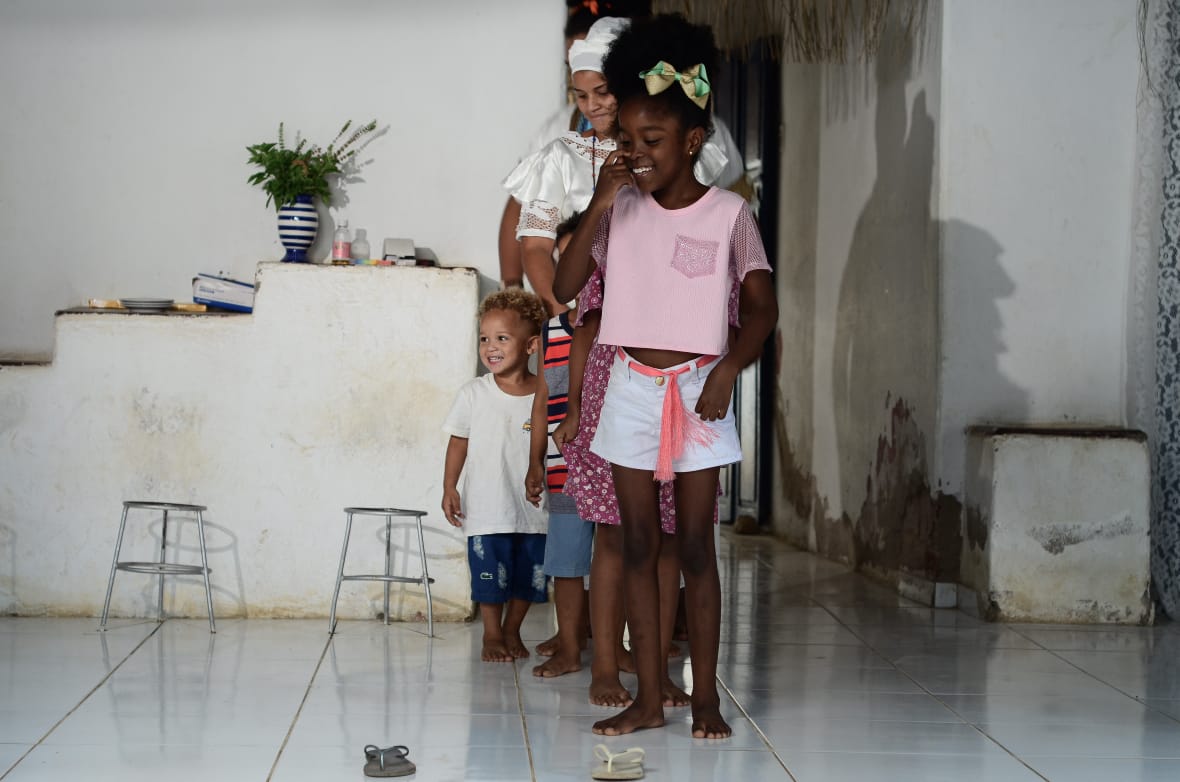 Uma menina negra com cabelos amarrados com um laço está sorrindo, com uma das mãos no rosto. Ela olha para o chão, onde há dois chinelos havaianas, um ao lado do outro. Atrás dela, há uma filha de outras crianças. 