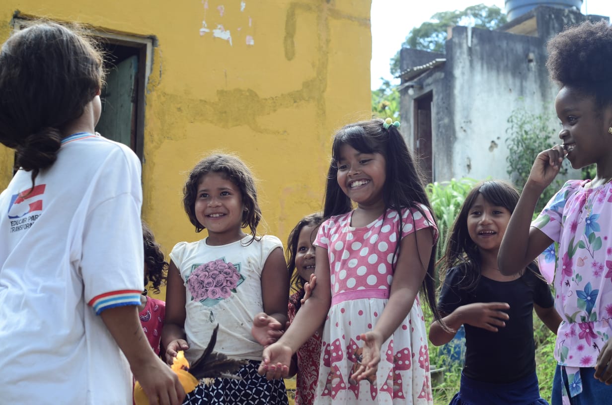  Uma senhora de cabelos brancos e roupa colorida segura uma peteca laranja nas mãos. Na frente dela, há cinco meninas, negras e com traços indígenas, que prestam atenção. 