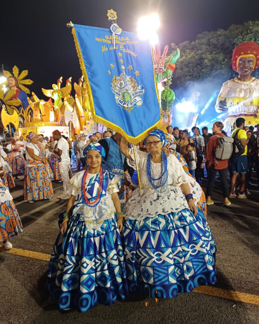 Imagem mostra avó e neta inspiração para o livro 'a menina que queria rodar a baiana'. Laura, uma menina vestida de baiana, e Bah, uma mulher idosa vestida de baiana segurando um estandarte.