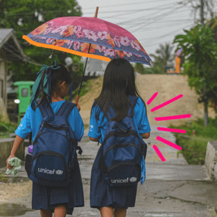 Imagem de capa para matéria sobre crise climática mostra dua smeninas de costas vestindo uniforme escolar, mochilas e uma delas segura um guarda-chuva. As duas atravessam uma ponte em uma rua de terra batida.