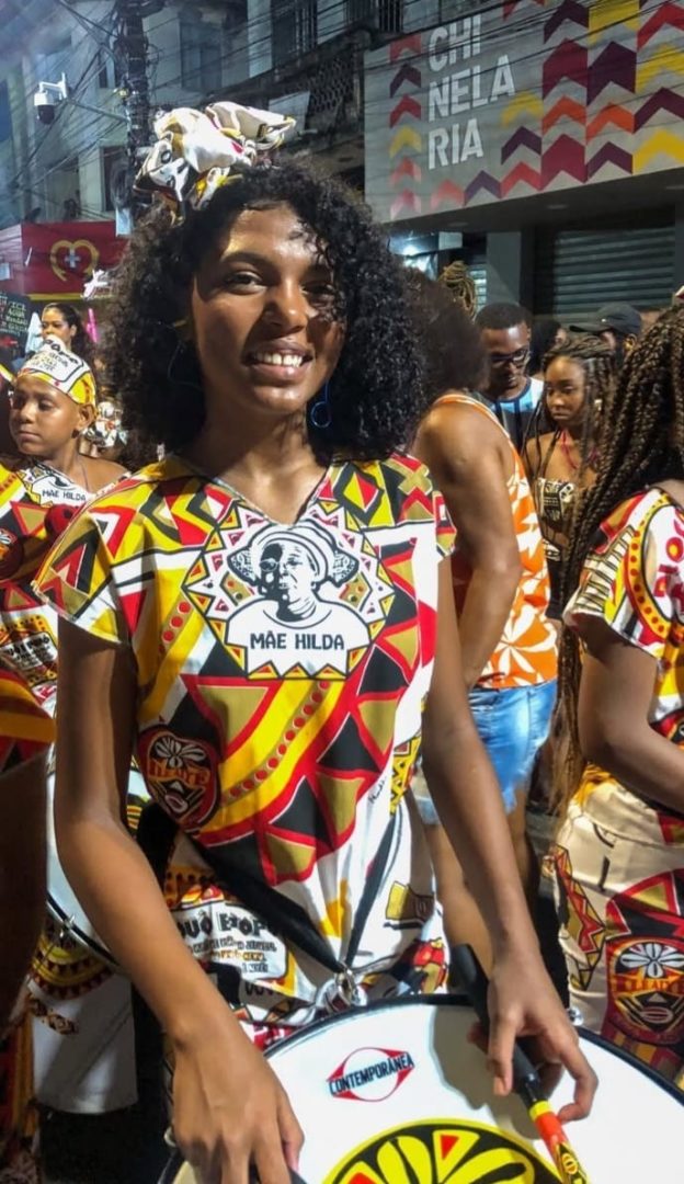 Imagem mostra uma menina negra, de cabelos cacheados, sorrindo e tocando tambor,