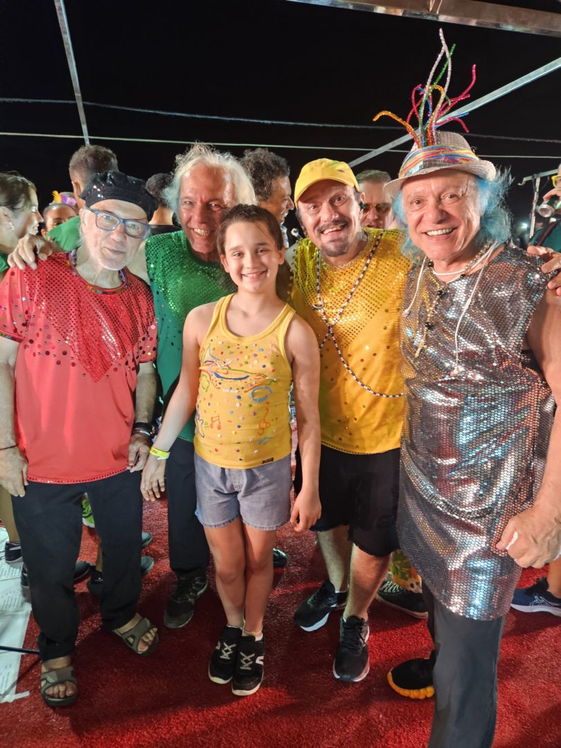 Imagem mostra quatro homens da família Macedo e Manu, uma menina branca, de cabelos lisos, sorrindo. Todos estão com roupas coloridas de carnaval. 