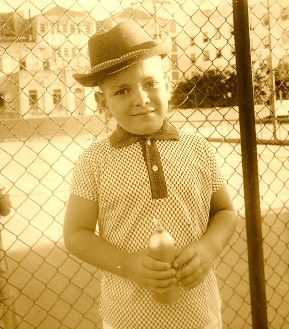 Imagem de foto antiga mostra um memnino usando roupa d carnaval e chapéu, segurando uma lata de metal. 