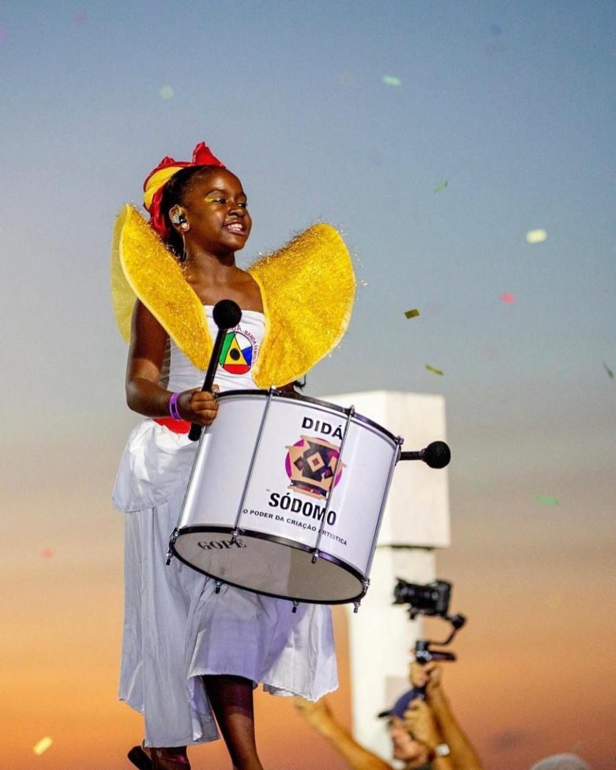 Imagem mostra uma menina negra, vestindo fantasia de bloco e tocando tambor.