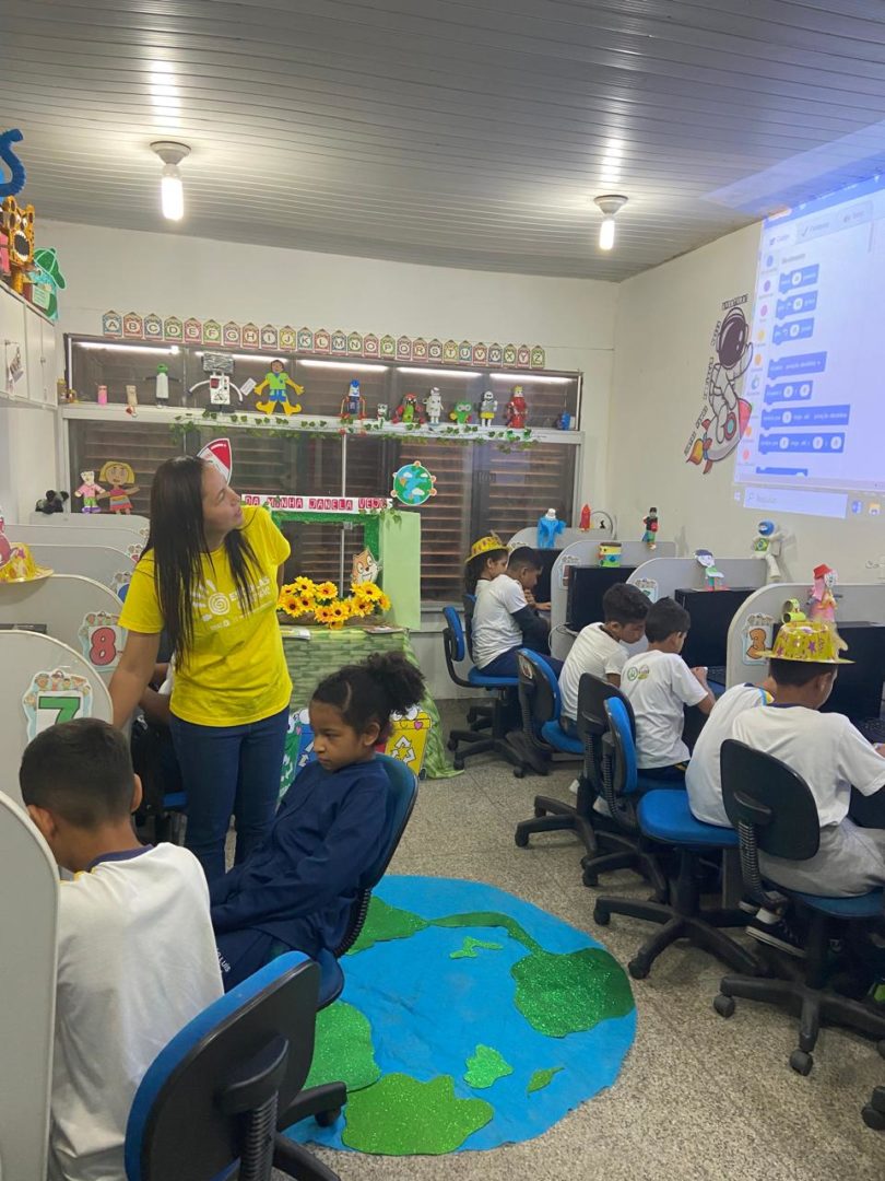 Imagem mostra uma sala de informática com uma professora ao centro e alunos sentados em frente a computadores.