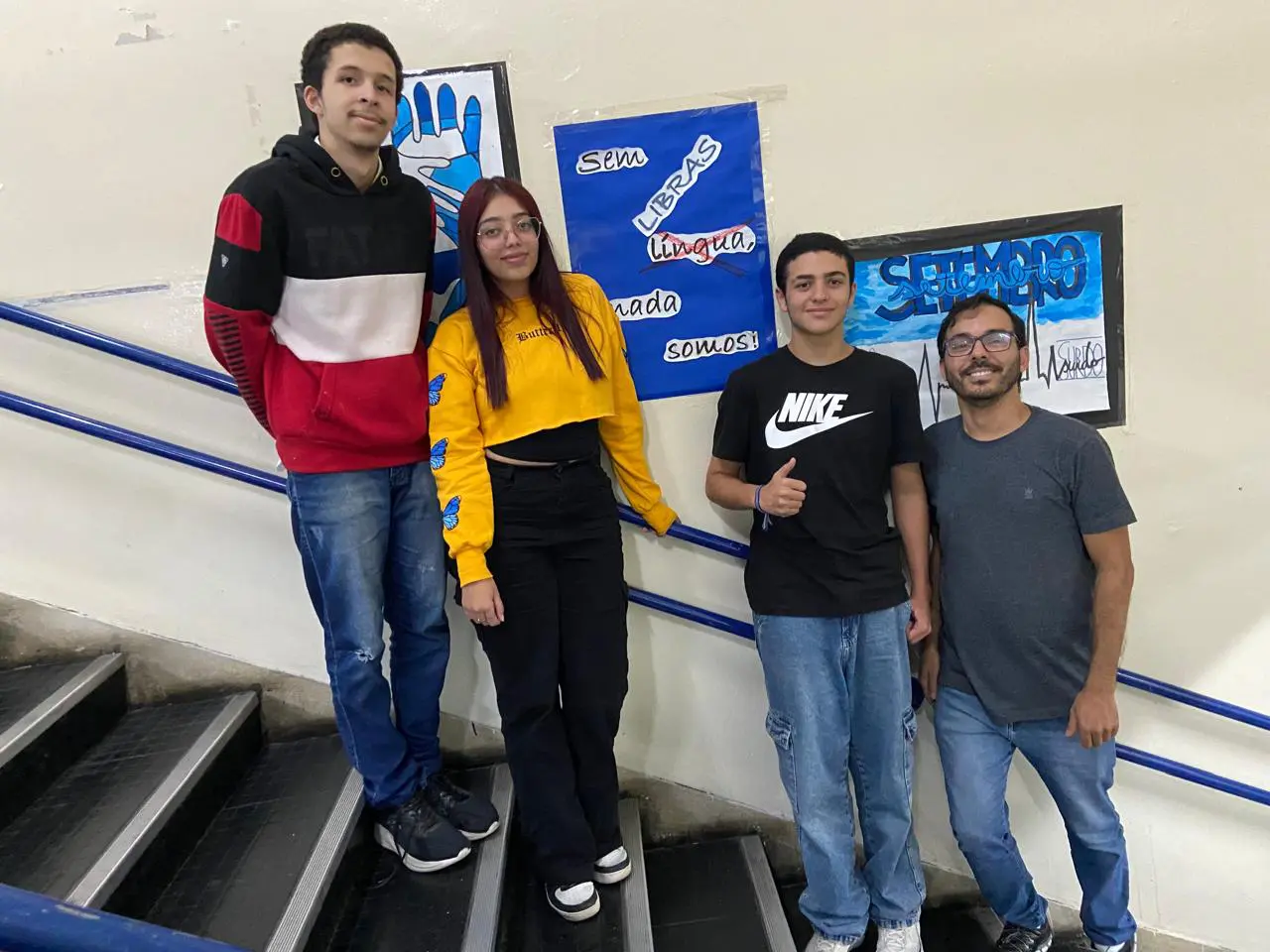 Imagem mostra alunos surdos e professor de Libras em corredor de escola. 