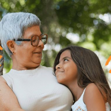 Foto de uma mulher de cabelos curtos e grisalhos abrançando uma menina de cabelos escuros. Ambas vestem roupas claras.