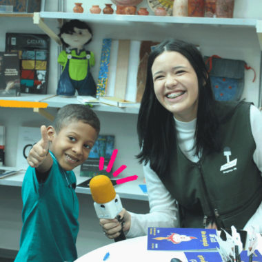 Imagem mostra adolescente de projeto de comunicação entrevistando criança com um microfone.