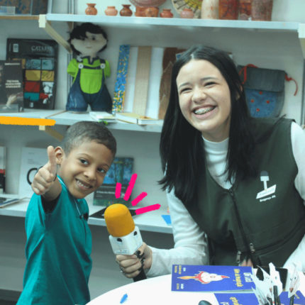 Imagem mostra adolescente de projeto de comunicação entrevistando criança com um microfone.