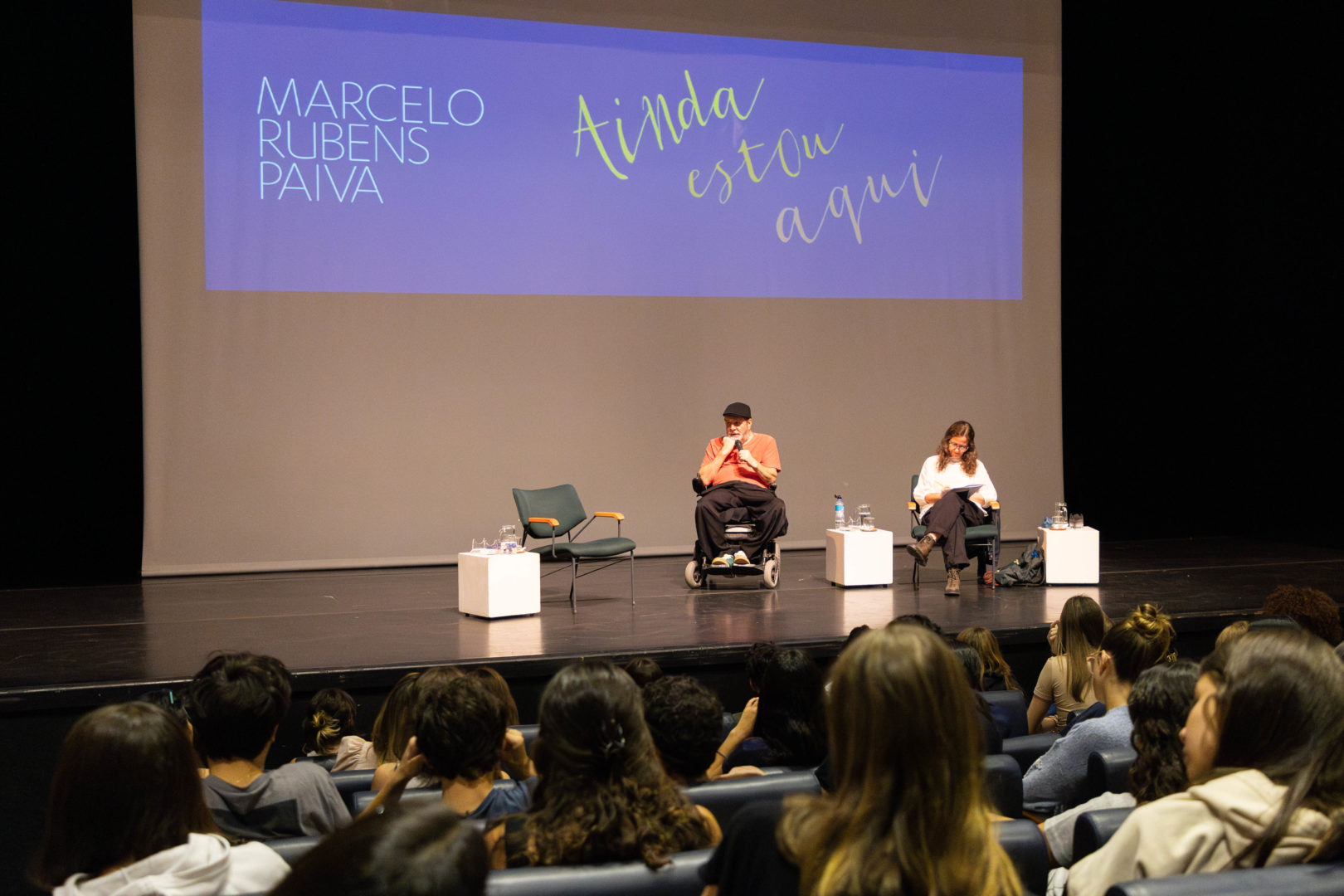 Imagem mostra o escritor do livro Ainda estou aqui, Marcelo Rubens Paiva, um homem branco, sentado em uma cadeira de rodas, falando em um palco de um auditório de escola.