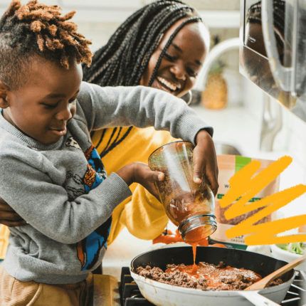 Uma mulher negra de tranças no cabelo e blusa amarelo ajuda um menino negro de blusa cinza a preparar uma comida na frigideira