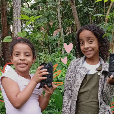 Imagem mostra duas crianças Xucuru de Ororubá: duas meninas negras de cabelos cacheados segurando mudas de plantas.