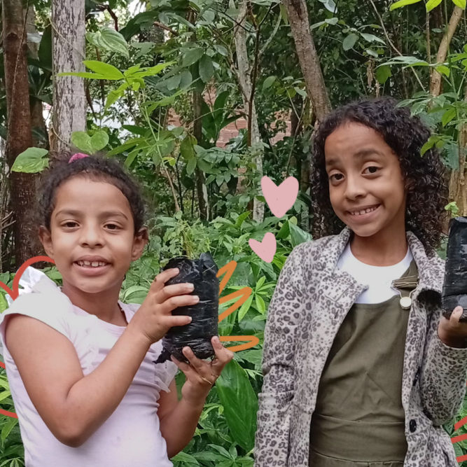 Imagem mostra duas crianças Xucuru de Ororubá: duas meninas negras de cabelos cacheados segurando mudas de plantas.
