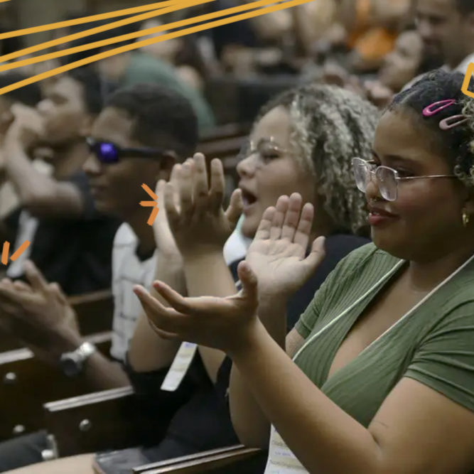 Imagem mostra um grupo de estudantes adolescentes negros sentados em um auditório e aplaudindo.