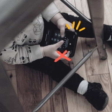 Imagem mostra uma criança pequena sentada debaixo de uma mesa e usando celular.