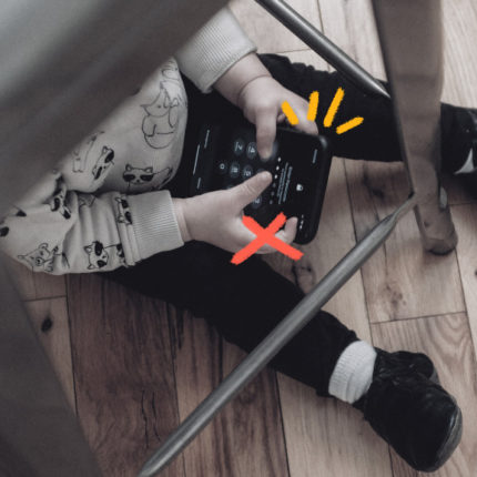 Imagem mostra uma criança pequena sentada debaixo de uma mesa e usando celular.