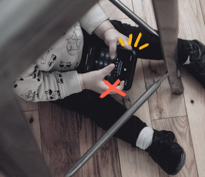 Imagem mostra uma criança pequena sentada debaixo de uma mesa e usando celular.