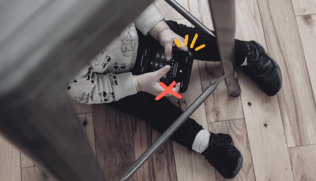 Imagem mostra uma criança pequena sentada debaixo de uma mesa e usando celular.