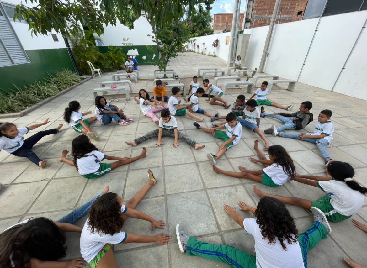 Imagem mostra uma turma de crianças fazendo alongamento em um pátio de escola.