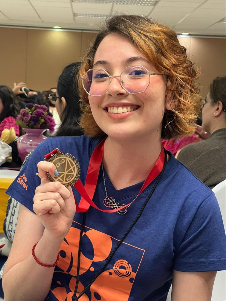 Imagem mostra uma adolescente de 15 anos, branca, cabelos curtos ruivos e óculos. Ela está sentada em um auditório e mostra uma medalha para a foto.