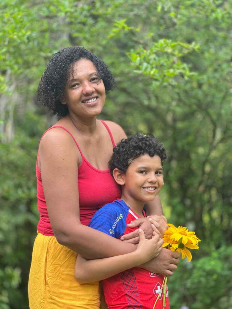 Imagem mostra Nara, uma mulher negra, de cabelos cacheados curtos e roupa vermelha. Ela abraça o filho Cristovam, um menino negro, de cabelos cacheados curtos.